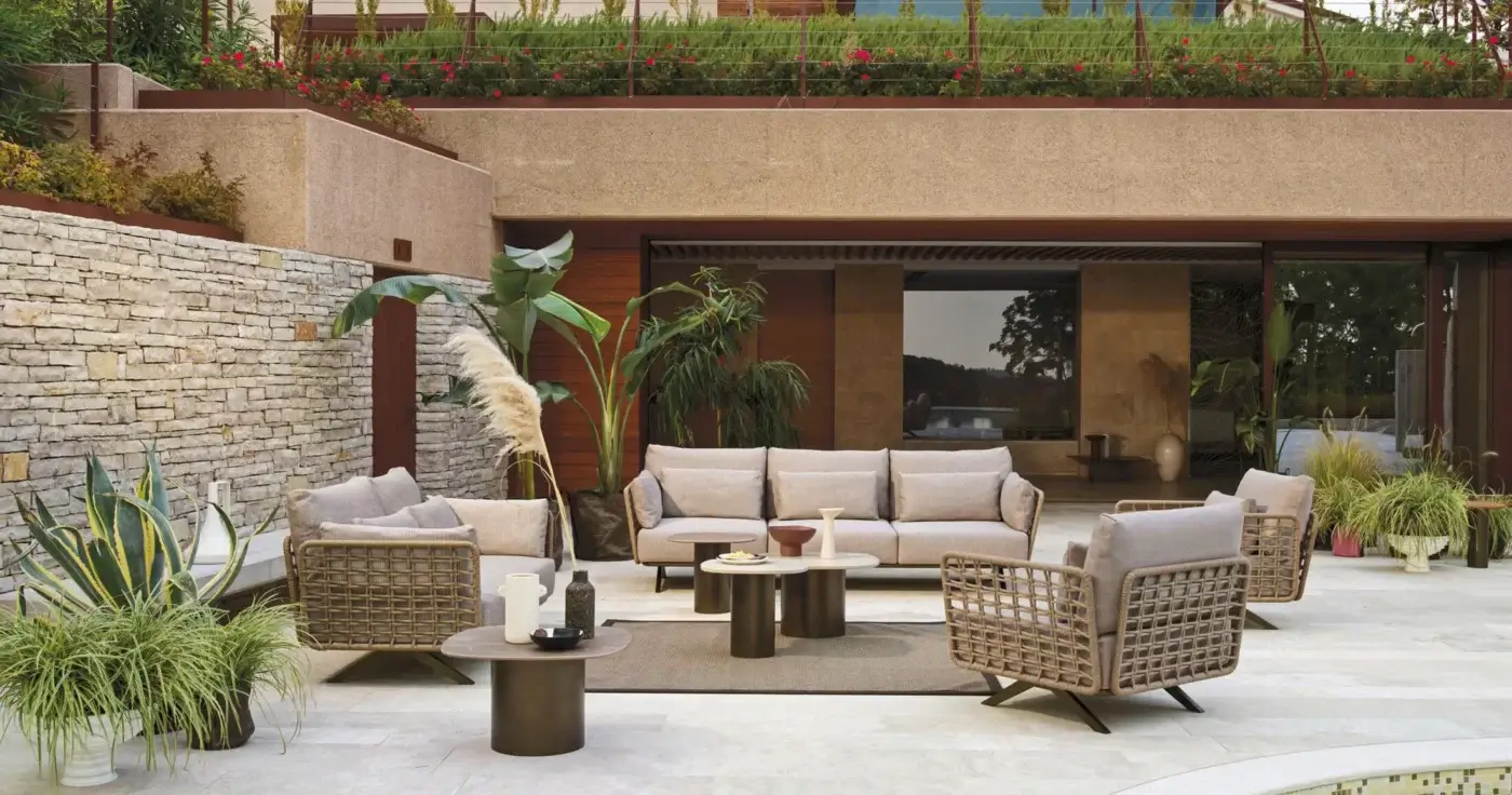 Rattan sectional sofa with light grey cushions on paved patio, flanked by matching armchairs and bronze side tables, modern outdoor setting.