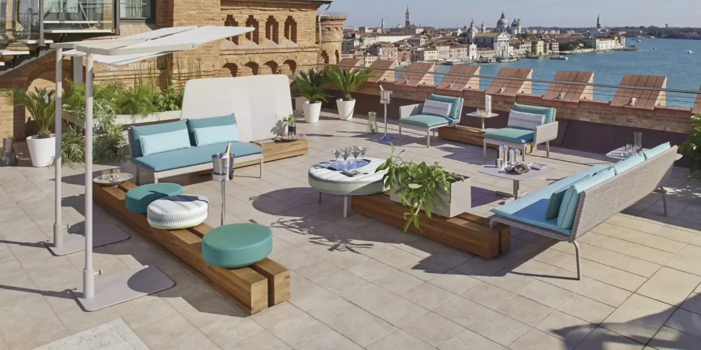 Rattan patio furniture set with turquoise cushions on a tiled rooftop terrace, featuring sofas, ottomans, and a white sculptural screen.