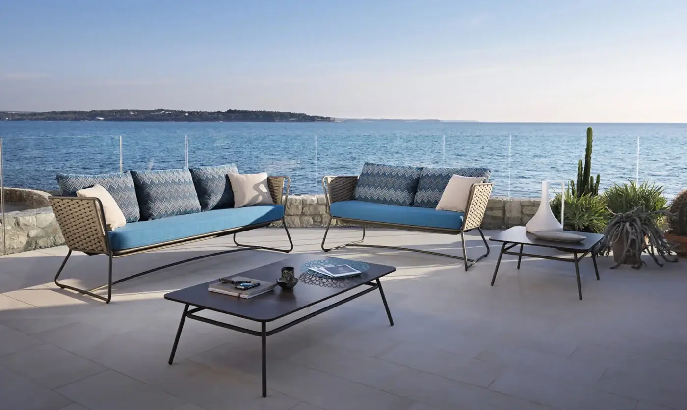 Roberti Rattan sectional sofa with blue zigzag fabric, dark metal frame, and geometric pillows on a tiled terrace overlooking water.