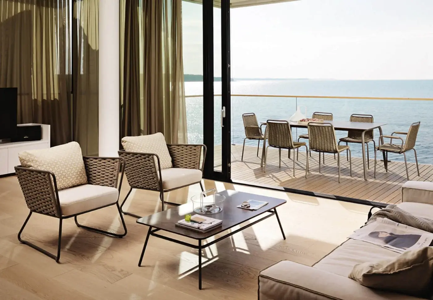Rattan seating area with beige cushions, dark wood coffee table, olive-green curtains, and water view through sliding glass doors.