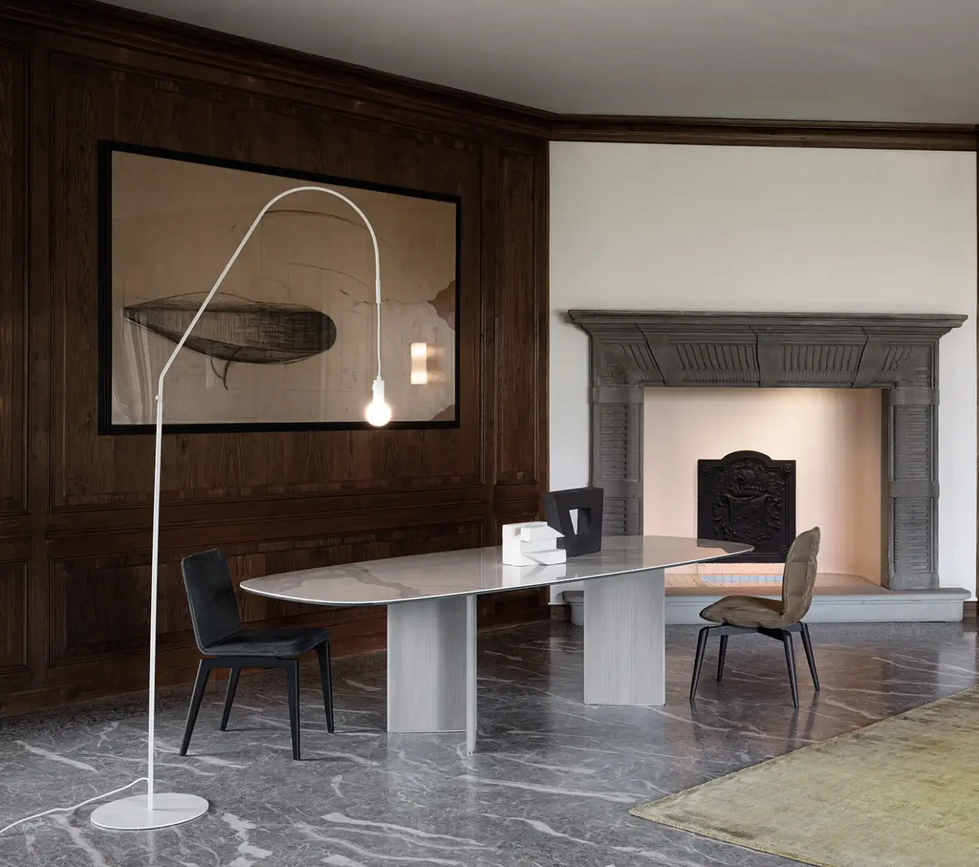 Dining table, white marble, elongated oval shape, with gray base and two chairs, in a room with wood paneling and stone fireplace.