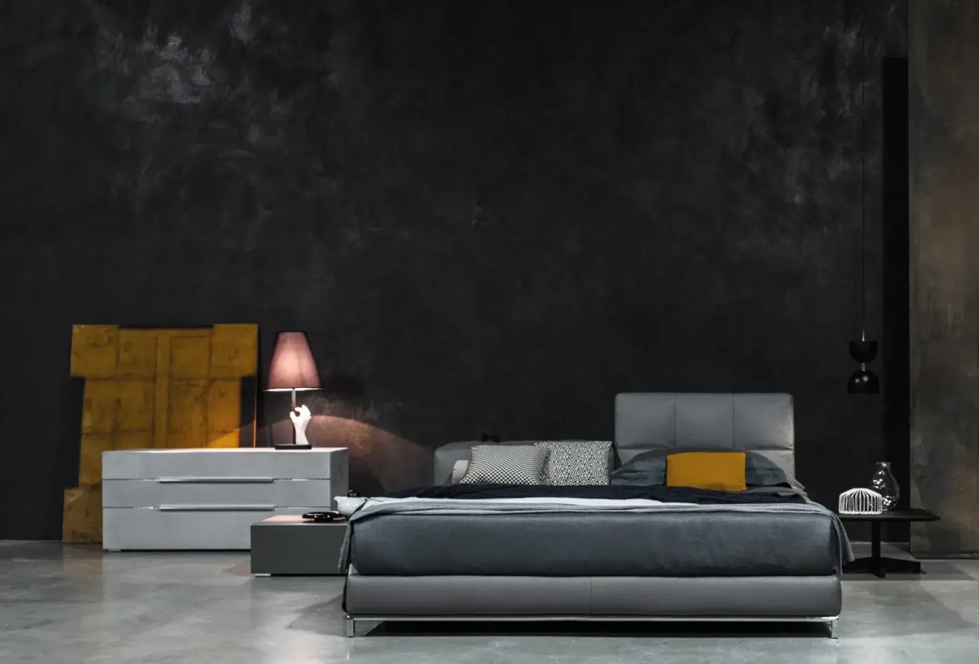 Gray leather platform bed with quilted headboard, mustard pillow, and geometric yellow dresser in a gray plaster bedroom scene.