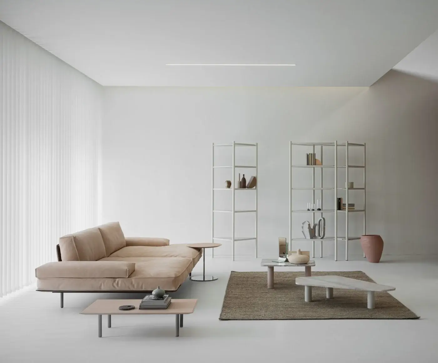 Beige leather modular sofa with low profile and wide armrests, flanked by white ladder shelving and round metal side tables in a minimalist living room.
