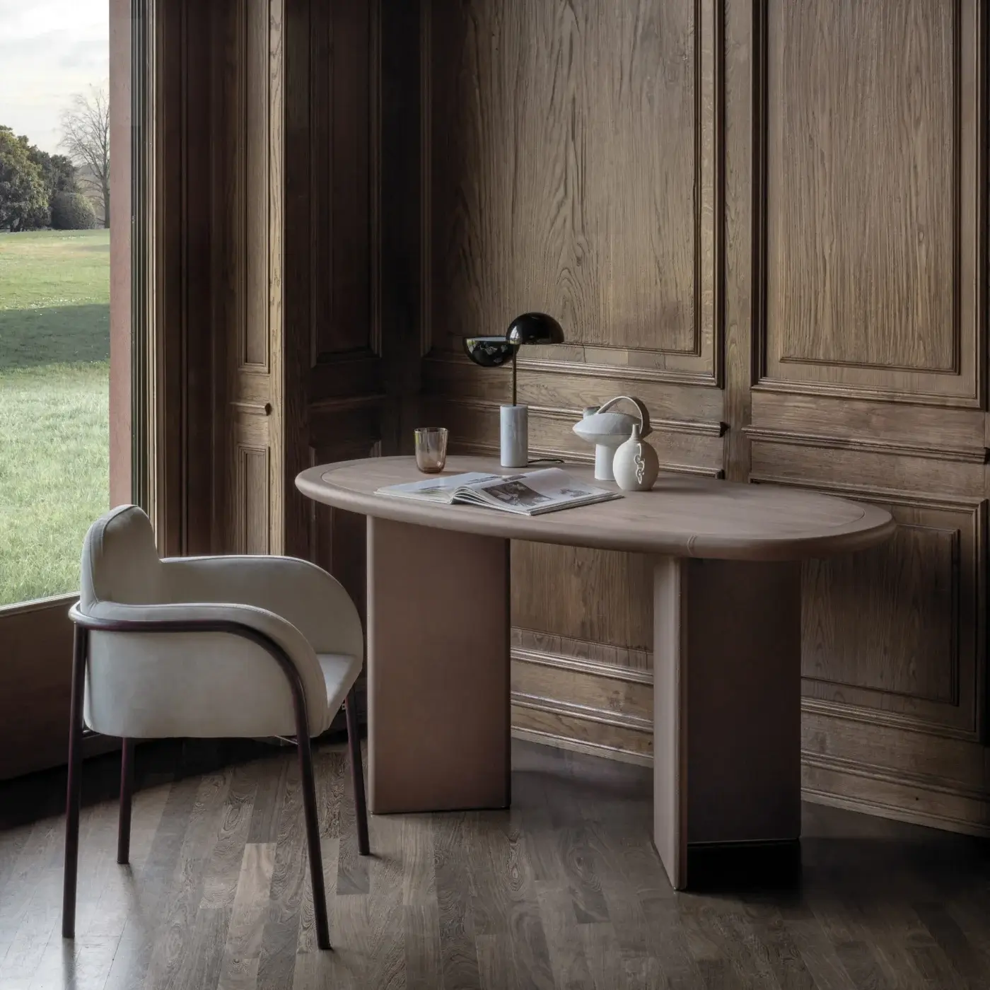 Zed desk, taupe finish, oval tabletop, with dark burgundy chair, bronze and ceramic lamps, in room with wood paneling.
