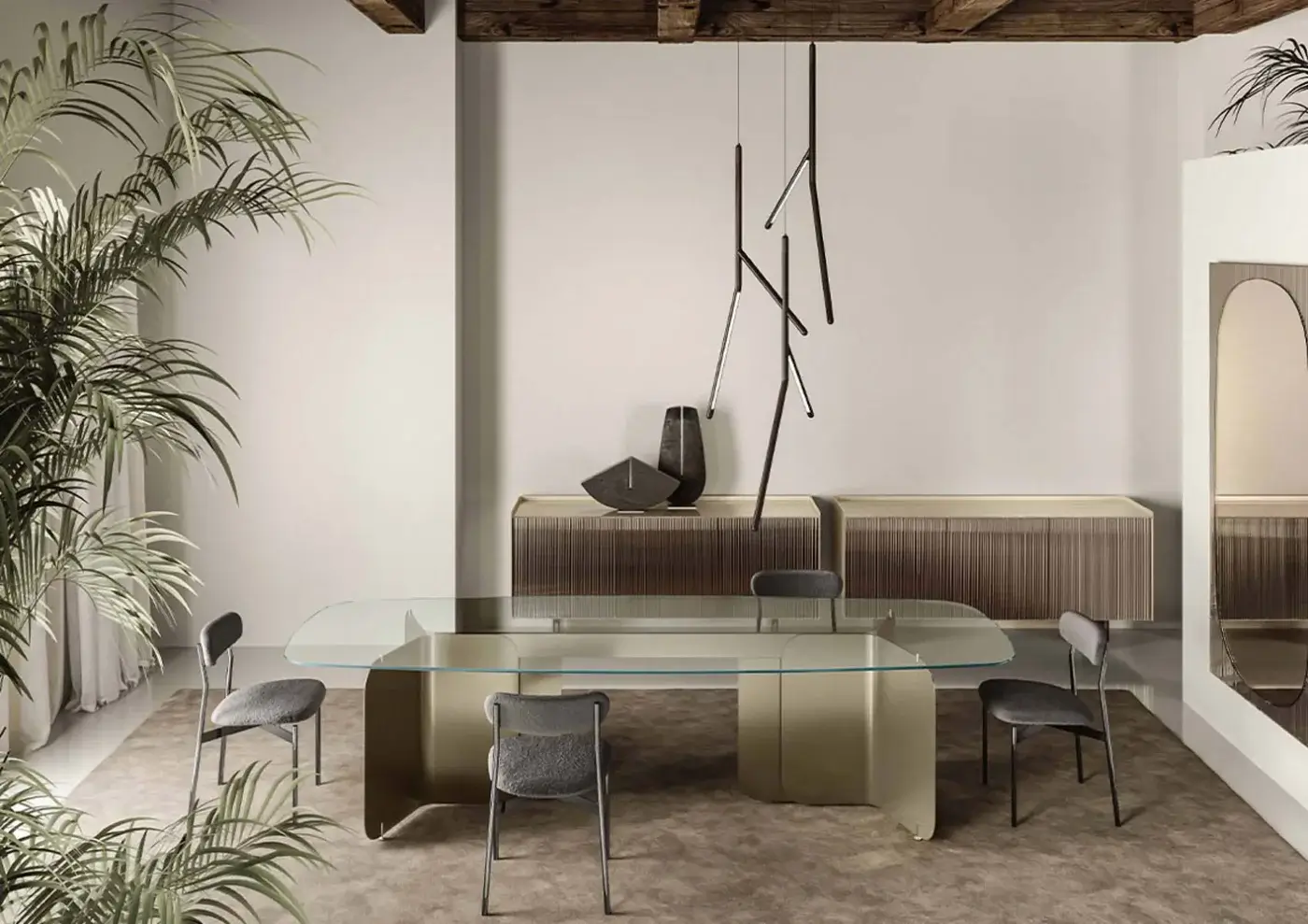 Clear glass dining table with champagne gold pleated base, surrounded by dark gray fabric chairs, in a beige room with palms.