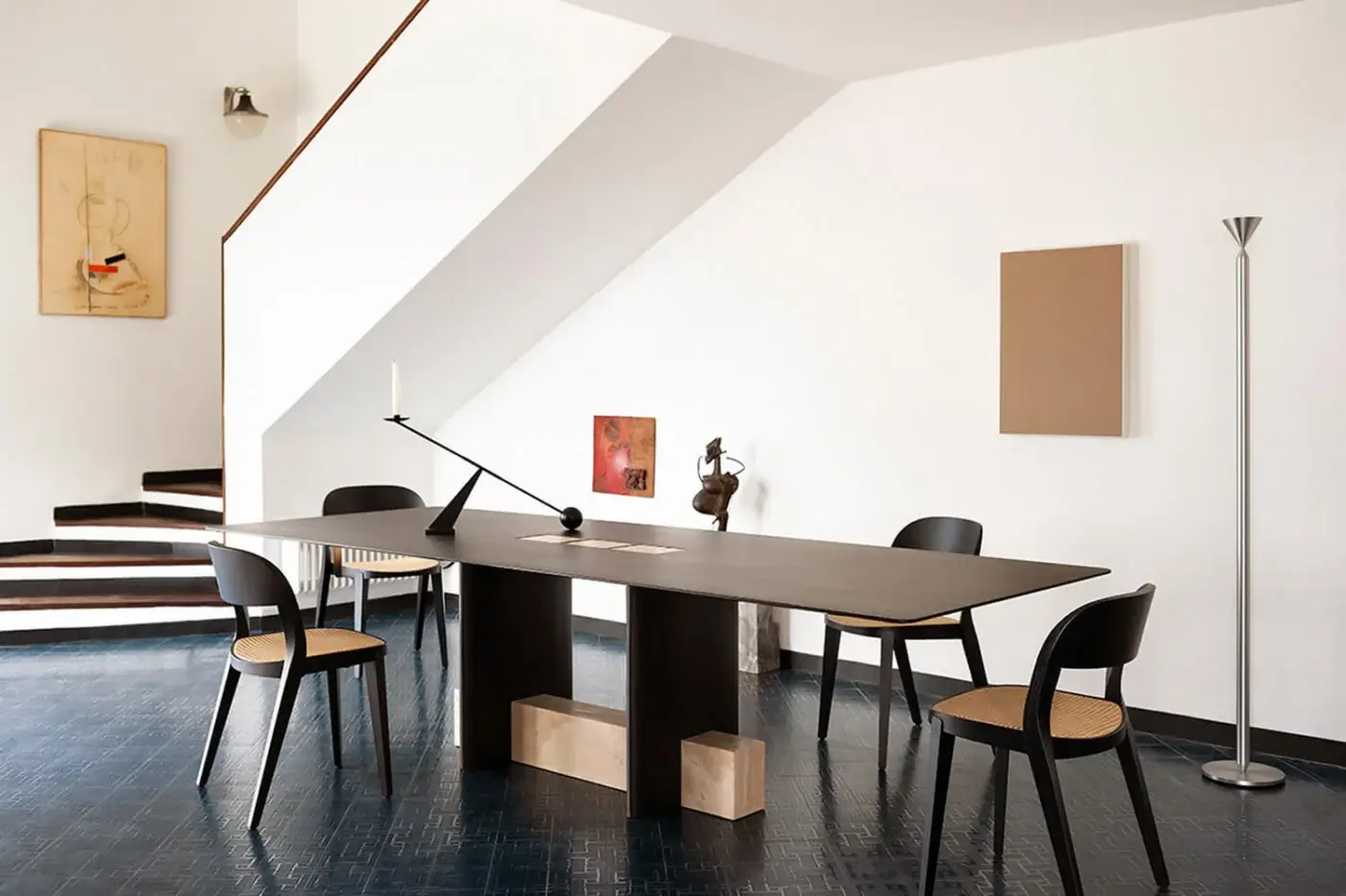 Dining table with dark gray stained wood top and three rectangular columns, surrounded by black chairs with woven seats, floor lamp, and artwork.