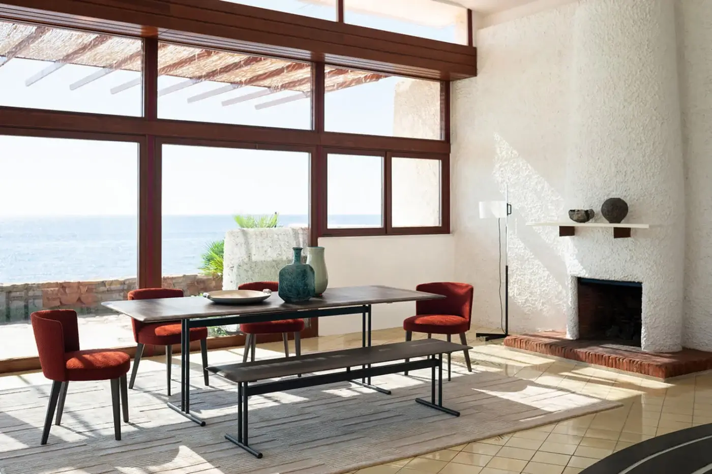 Dining table with stained wood top and metal legs, surrounded by four red chairs, bench, ocean view, stone fireplace, white walls.