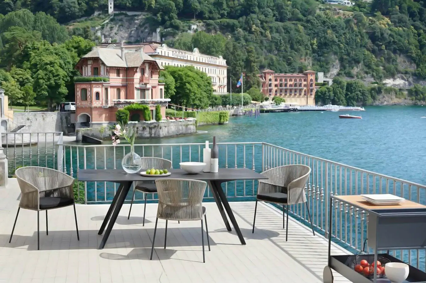 Potocco Katans table, dark gray circular top, black angled metal base, with four woven light beige chairs, lakeside terrace setting.