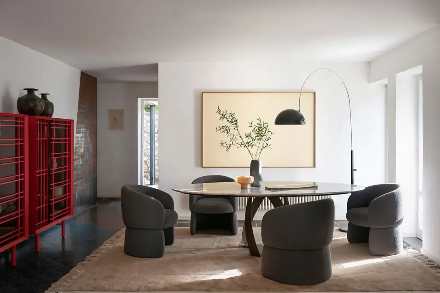 Dining room with round marble table, gray bulbous chairs, black arc lamp, botanical artwork, and red Potocco "Sen" cupboard with ceramic vases.