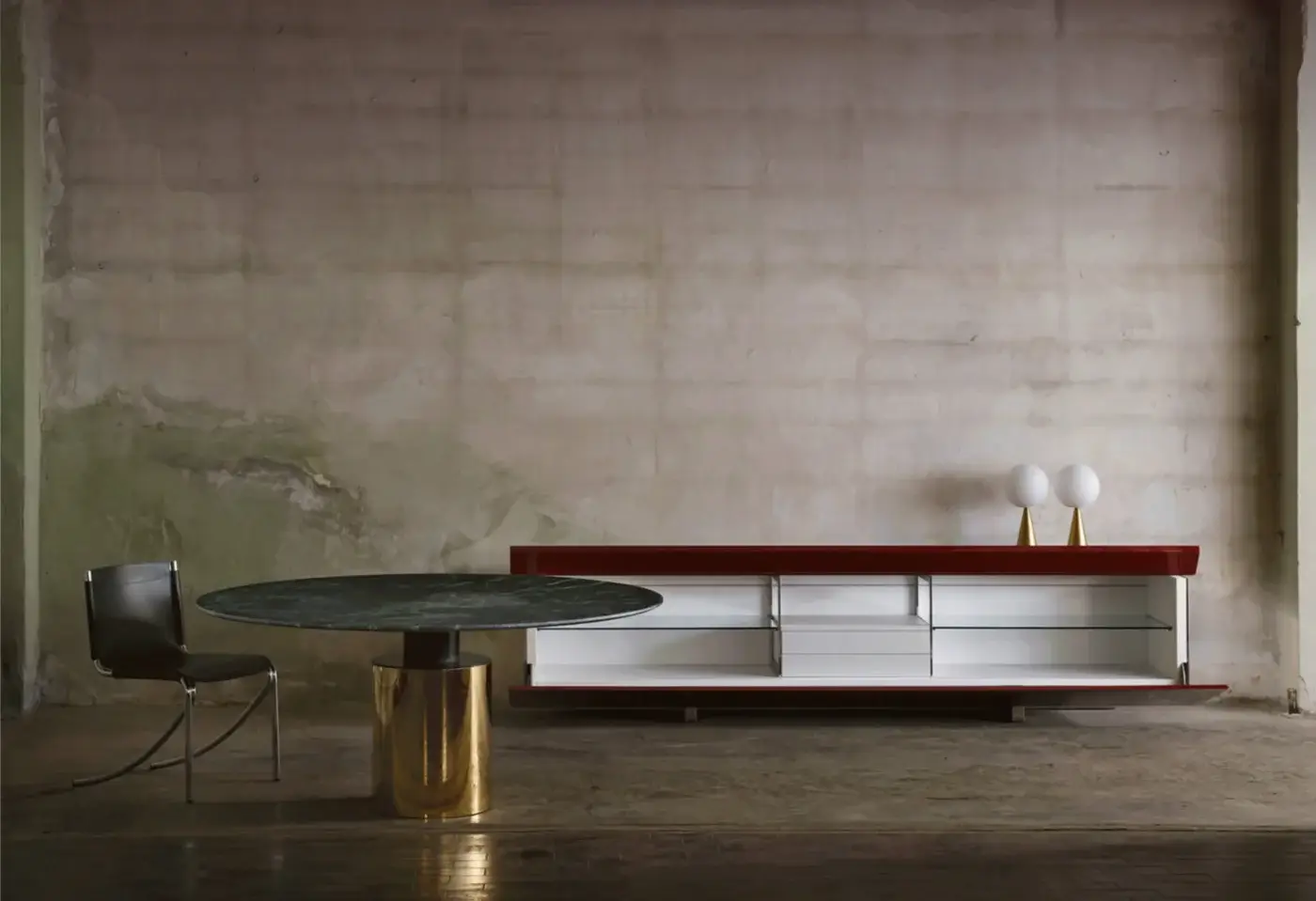 Round Creso table with dark green marble top and brass cylindrical base, beside a red console with white drawers.
