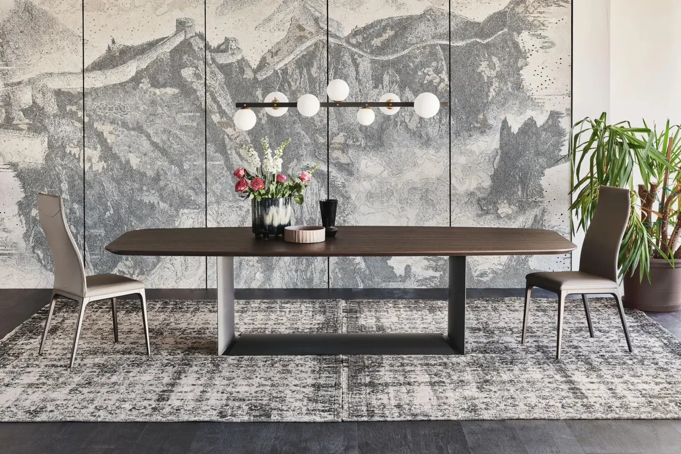Dragon Wood dining table, dark brown wood, gray metal base, with floral centerpiece, linear chandelier, and grayscale mountain backdrop.