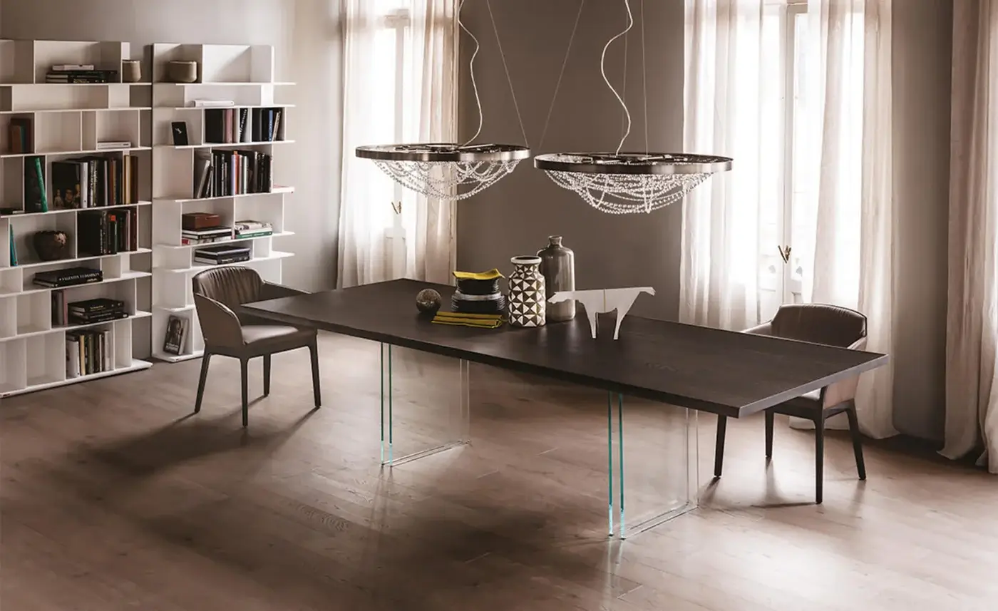 Ikon dining table with dark wood top and glass X-base, surrounded by eight light brown leather chairs, under crystal pendant lights.