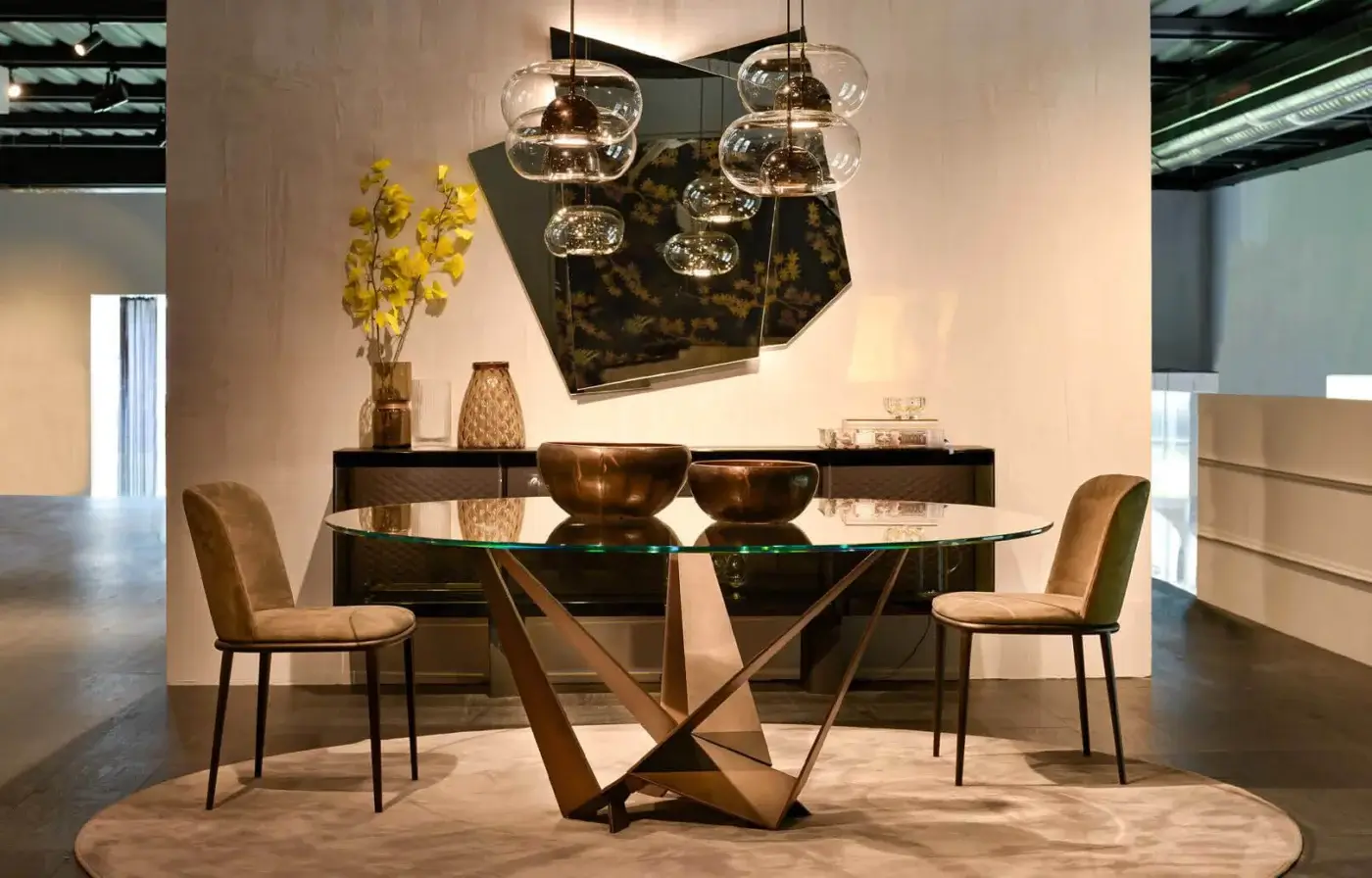 Skorpio dining table, clear glass top, bronze metal starburst base, paired with gray fabric chairs, on a beige rug in a modern dining room.