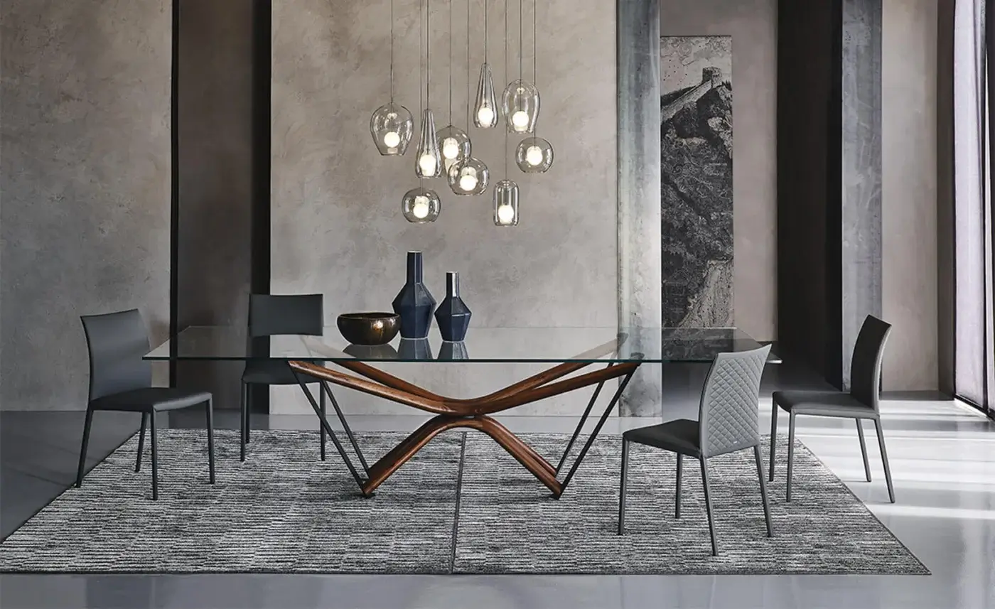 Glass dining table with dark wood wing-shaped base and six gray quilted chairs on a textured rug, lit by glass pendant lights.