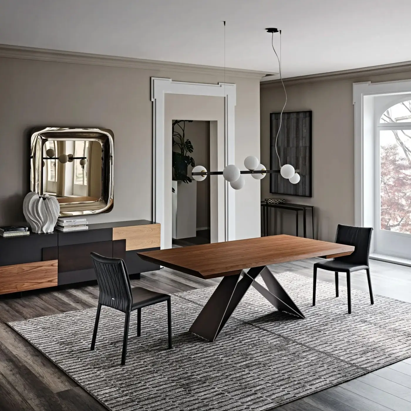 Dining table, wood and metal, rectangular with triangular base; gray chairs, sideboard, chandelier, mirror, and patterned rug in a modern dining room.