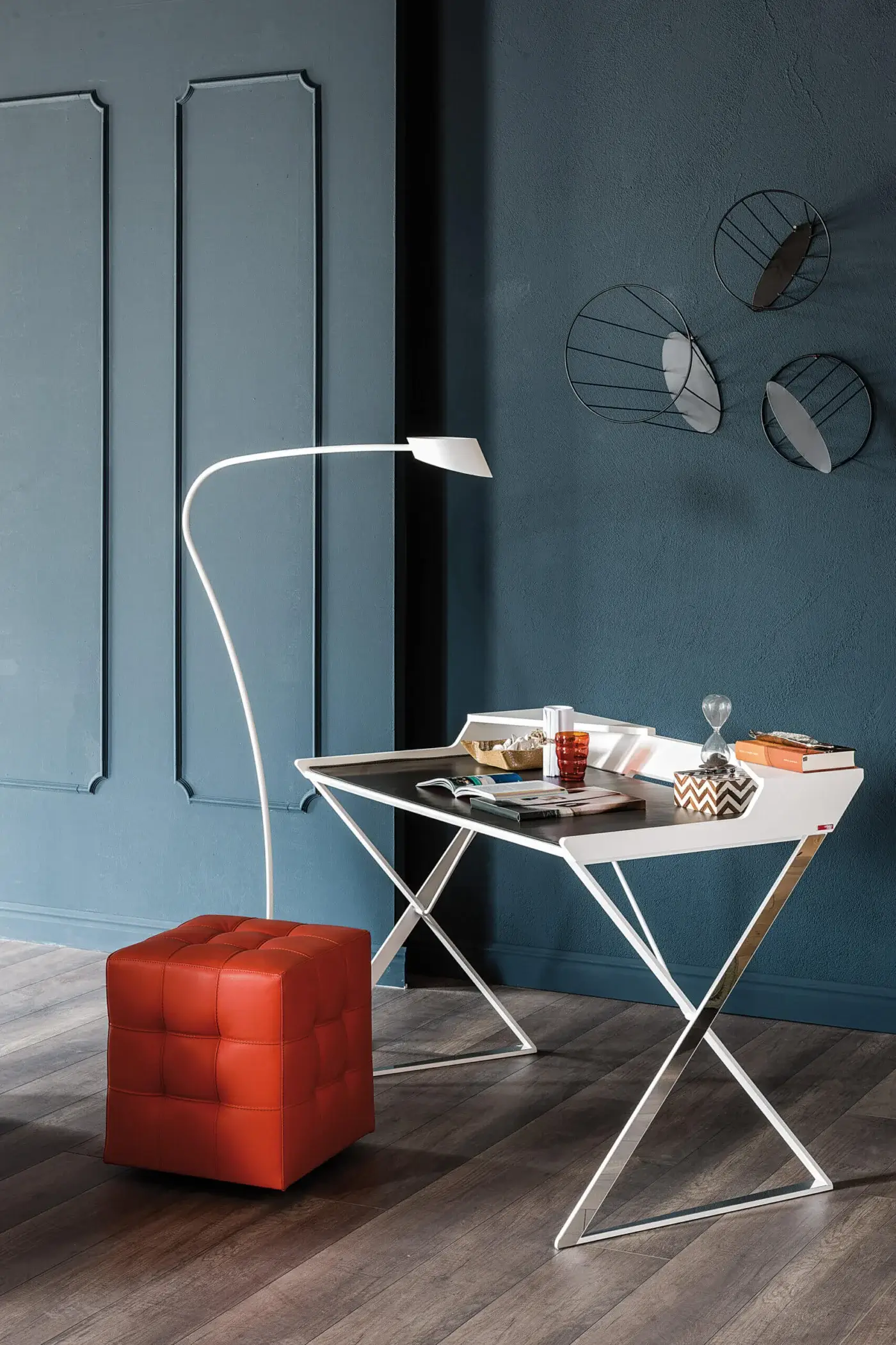 Cattelan Italia qwerty desk, white tabletop, chrome X-leg, teal wall, orange leather ottoman, arc lamp, modern minimalist office setup.