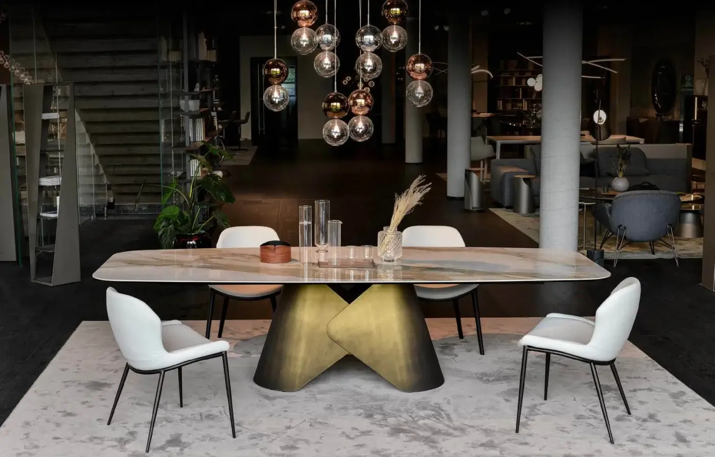 Ceramic table with white marble-effect top and burnished bronze metal inverted cone base, surrounded by six white upholstered chairs.