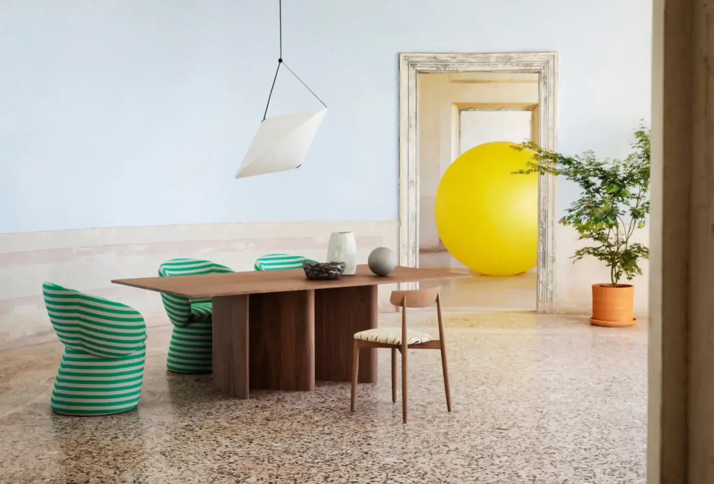 Nami dining table, wood grain top, geometric base, paired with striped armchairs, pendant light, terrazzo floor, and yellow sphere backdrop.