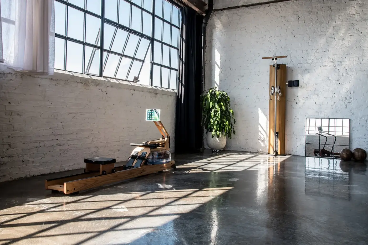 Rowing machine with light wood frame and dark seat, next to a wooden Pilates reformer, in a bright studio with white brick walls.