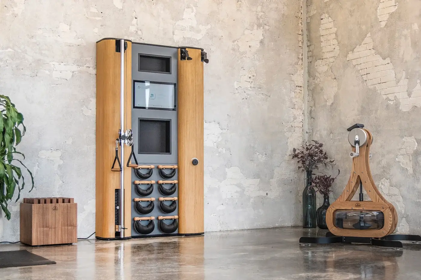 Fitness cabinet with metal bar and resistance bands, Waterrower rowing machine, beige brick wall, concrete floor.