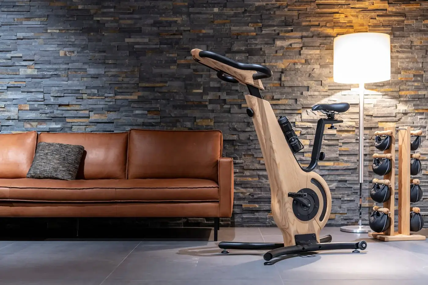 Waterrower Nohrd exercise bike with light wood frame and black metal accents, positioned in a modern living room with stone wall.