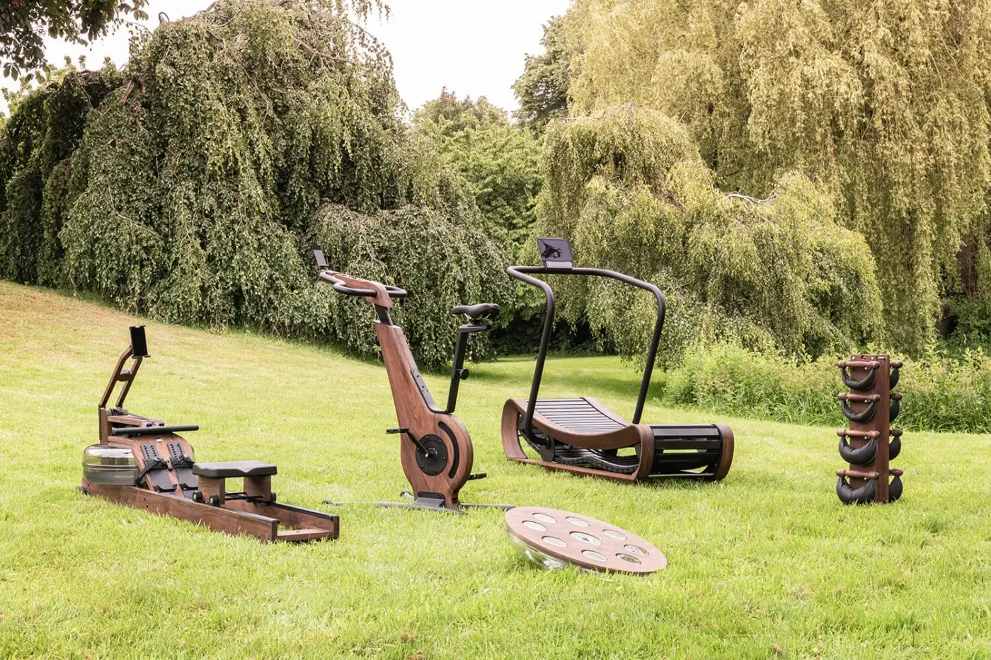 Waterrower Nohrd fitness equipment on lawn: rowing machine, bike, trainer with dark wood frames, black metal, weighted discs, natural lighting.