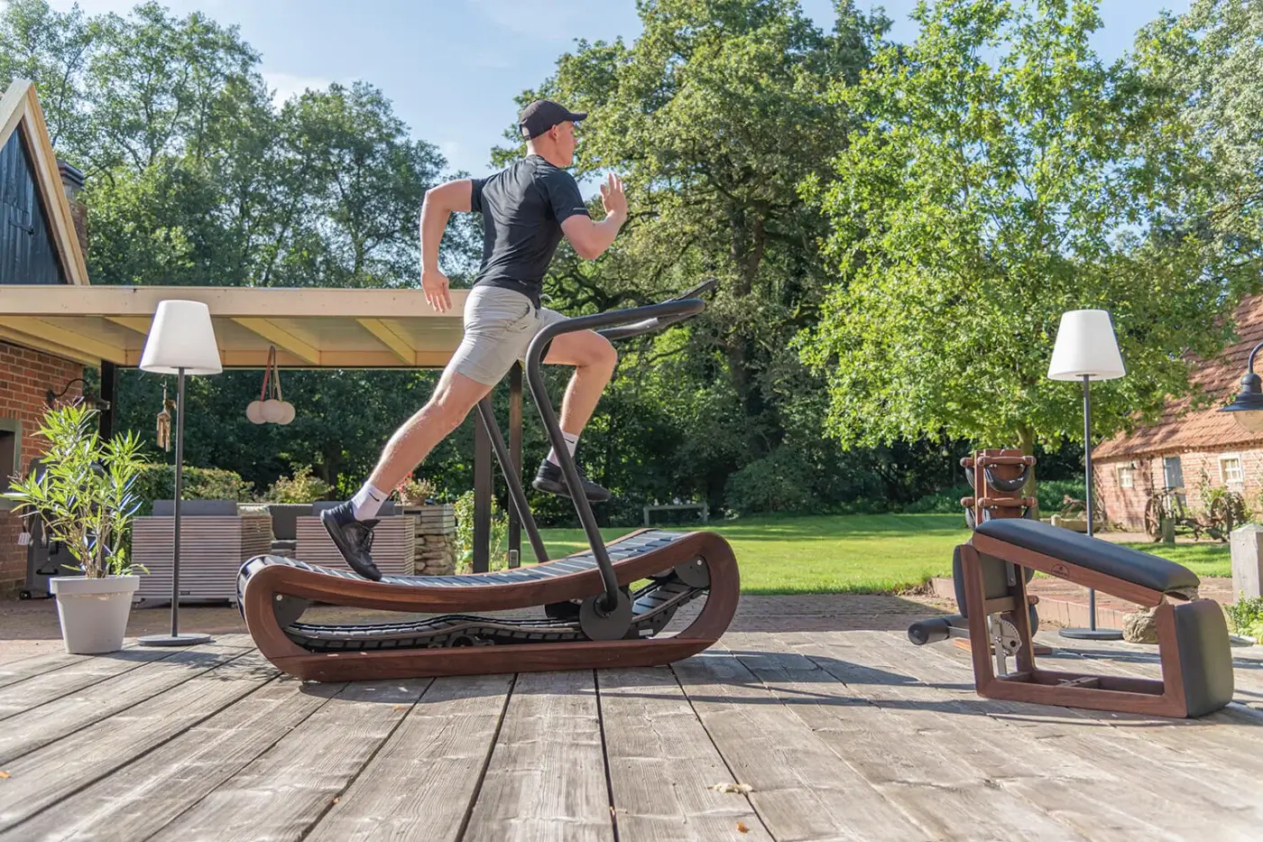 WaterRower Nohrd fitness machine with dark wood frame and black belt on a wooden deck, with bench, lamps, and greenery.