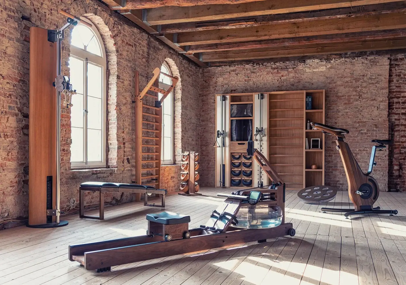 Waterrower Nohrd rowing machine with glass tank on wood floor in home gym with pull-up station, dumbbells, and exercise bike.