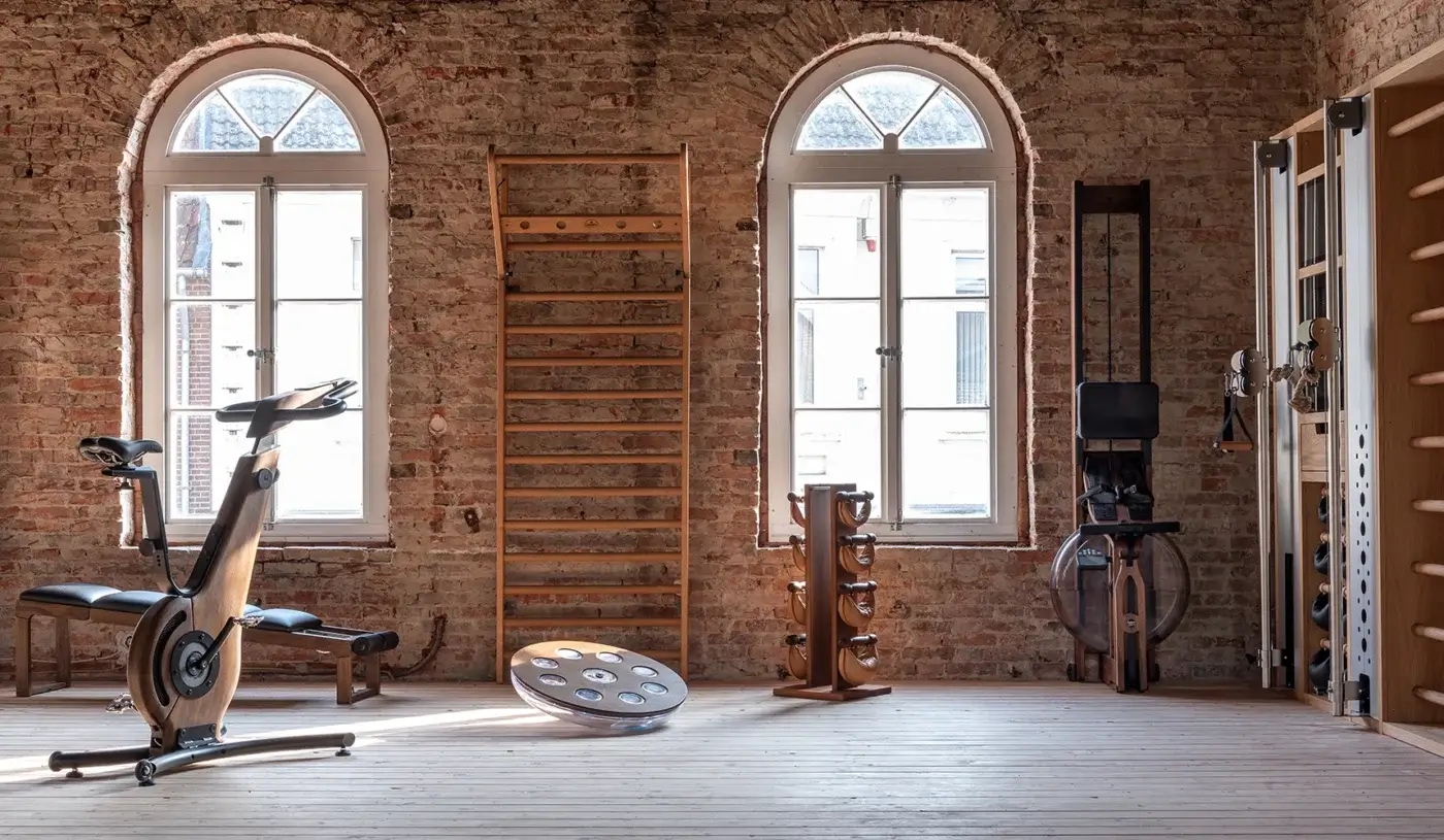 Home gym interior with Waterrower rowing machines, wooden Swedish ladder, dumbbells, and balance disc against exposed brick walls.