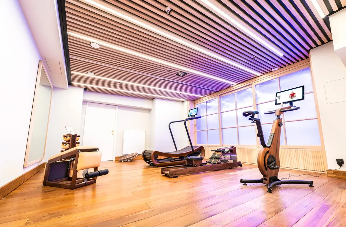 Waterrower Nohrd rowing machine on honey hardwood floor, dark wood slat ceiling with LED lights, treadmill, strength station.