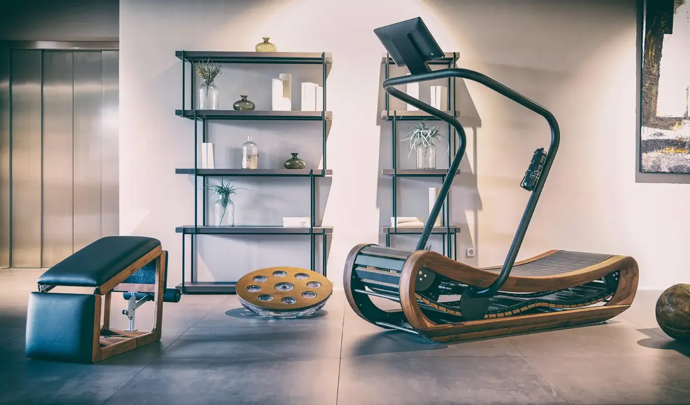 Waterrower Nohrd fitness machine with curved wooden base and black metal frame, in a minimalist gray tiled interior with bench and shelving.