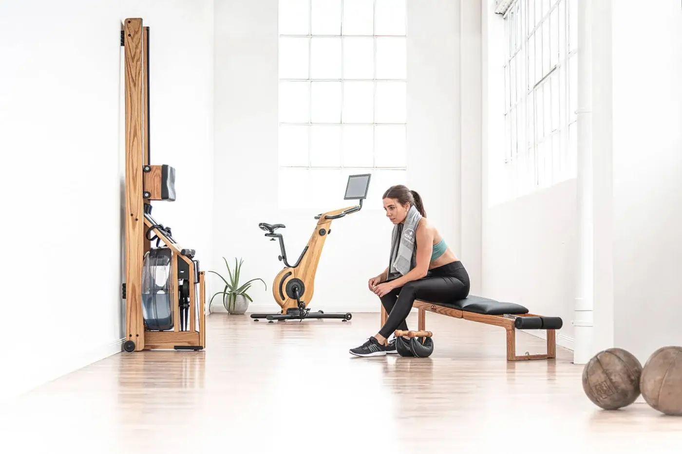 Waterrower Nohrd exercise machine with wooden frame, a training bench, medicine balls, and a woman exercising in a bright studio.