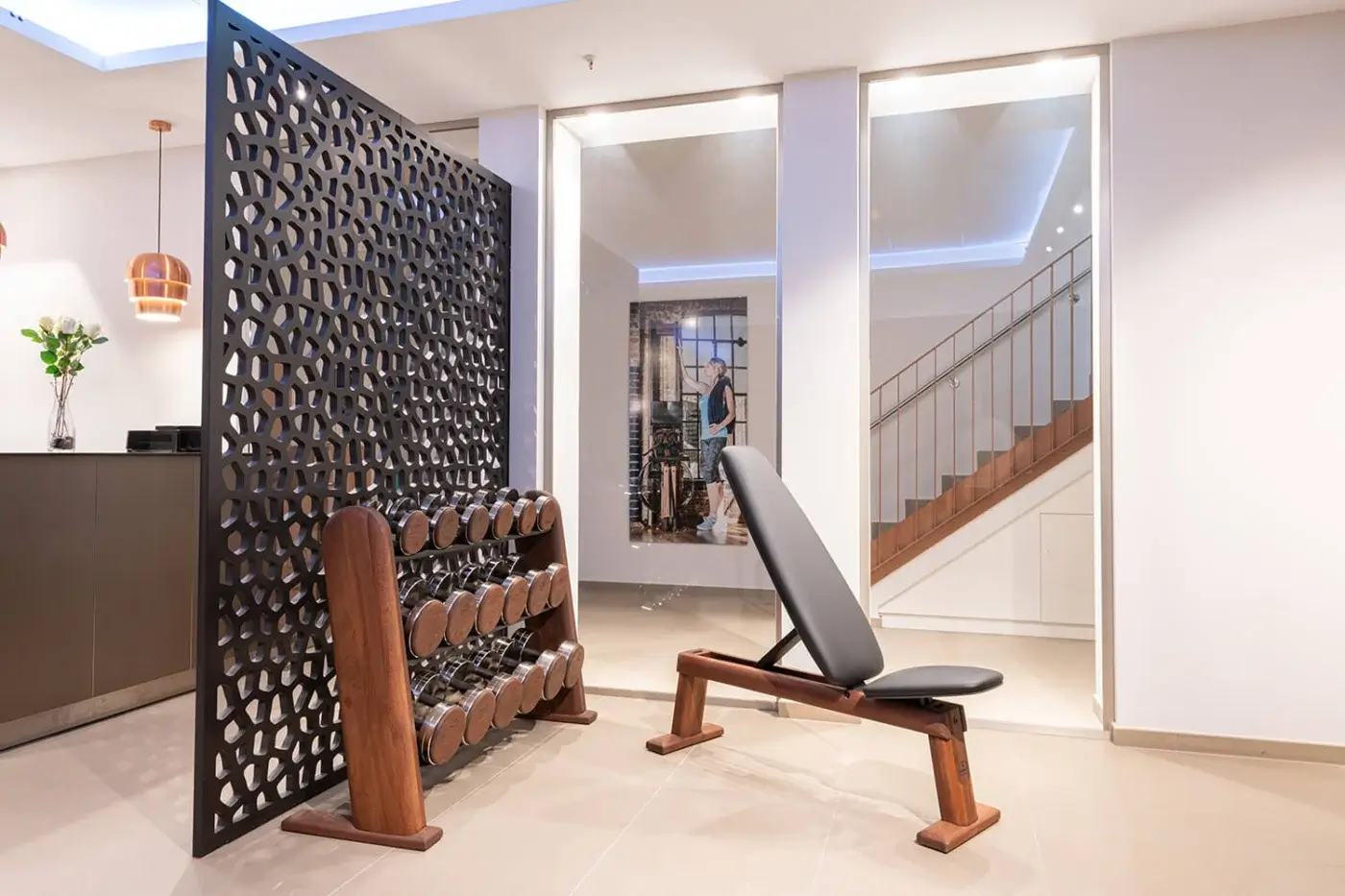 Home gym with dark wood dumbbell rack, weight bench, geometric screen, copper pendant light, white staircase, and mirrored wall reflecting interior.