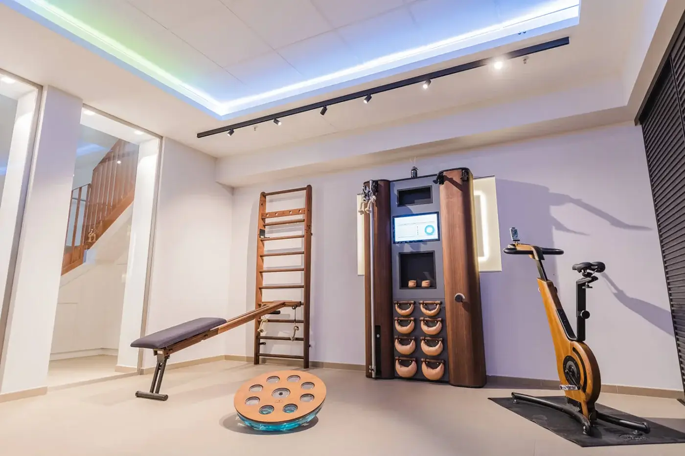 Home gym interior with Waterrower rowing machine, wood slatted wall, weight rack, Swedish ladder, balance board on beige tile floor.
