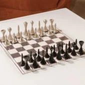 Chess table, silver metal and dark gray board, with conical pieces and a brushed finish, set against a white surface and brown leather sofa.