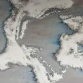 Cloudscape aerial view, white and gray tones, dense textured formations follow land contours, revealing glimpses of blue sky and coastline.