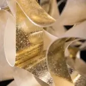 Layered paper ribbons, off-white and gold leaf, display a textured, crackled pattern with overlapping curves and a flowing, organic structure against black.