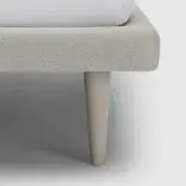 Bed frame detail, light gray herringbone upholstery, cylindrical beige leg, white fitted sheet, plain white background, solid material construction.