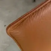 Leather chair corner, tan-brown full-grain leather, rounded shape with dark brown trim and detailed stitching on neutral gray concrete.
