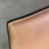 Leather cardholder detail, dark brown and tan, with visible yellow hand-stitching along a raw, layered edge and natural grain texture.