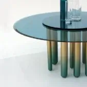 Round coffee table, blue glass top, cylindrical legs with sapphire to gold gradient finish, studio setting.