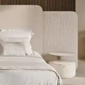 Upholstered bed with cream embroidered headboard, white linen bedding, and circular white bedside table with silver legs.