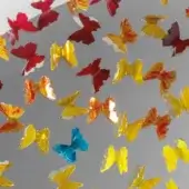 Metallic butterfly decorations in yellow, orange, red, and blue hang from a gray reflective surface, creating distorted reflections and a dynamic composition.