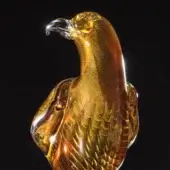 Glass falcon sculpture, amber and gold, with swirling patterns and a streamlined profile, shown against a black backdrop.