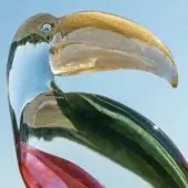 Glass toucan sculpture with gold beak, clear body, and green-red accents, displayed against a pale blue backdrop.