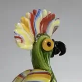 Glass parrot sculpture, translucent green with yellow and red chest, black beak, and colorful swirling feather crest detail.