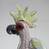 Glass cockatoo sculpture, pink with purple, features a textured green crest and translucent glass strand against a gray backdrop.