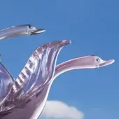 Glass swan sculptures, violet and pink, depicted in flight with curved wings against a clear blue sky and single white cloud.