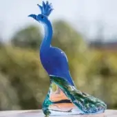 Blue glass peacock sculpture with translucent body, swirling green and gold tail feathers, and clear, tinted legs on a reflective surface.