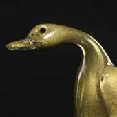 Glass duck sculpture, gold translucent finish, stylized form with curved neck and black bead eye against a black background.